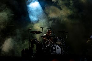 A drummer performs on stage with dramatic blue-tinted lighting and smoke effects during a concert performance.