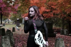 Gothic figure in black dress poses in autumn cemetery with jack o'lantern guitar among red maple leaves and gravestones.