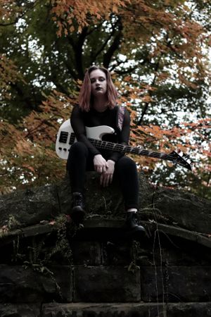 Moody portrait of musician with bass guitar sitting on stone wall against autumn foliage.