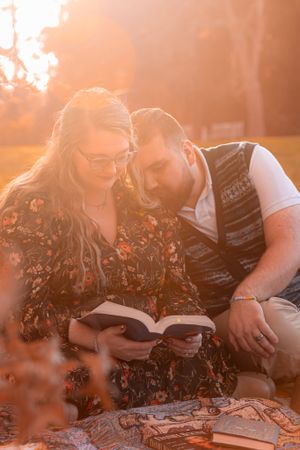 A couple reads a book together on a blanket during golden hour sunset with warm orange lighting filtering through trees.