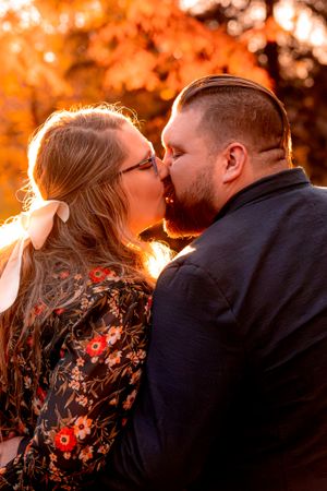 A couple shares a romantic kiss in front of vibrant orange autumn foliage at sunset.