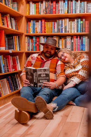 A cozy scene of two people reading together on the floor against wooden bookshelves filled with colorful books.