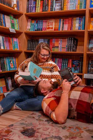 Two readers relax with books on a cozy couch in front of wooden bookshelves filled with colorful novels at a library or bookstore.