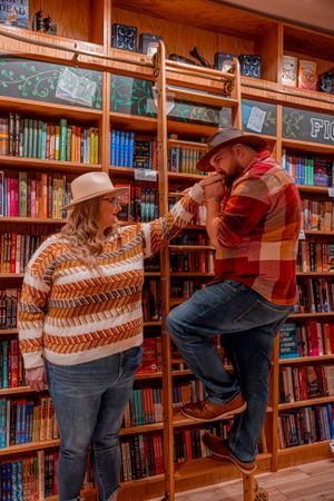 Two people in cozy sweaters interact playfully by wooden library bookshelves filled with colorful books.
