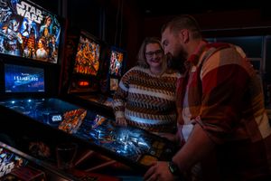 Two people play pinball at an arcade with Star Wars themed games and lighting in the background.