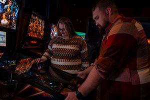 Two people play a vintage pinball machine in a dimly lit arcade with neon signs glowing in the background.