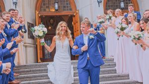 Wedding party celebrating on church steps with bride and groom in blue suit surrounded by bridesmaids in lavender dresses.