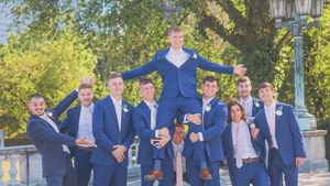 Groomsmen in navy suits lift and balance a figure in a blue outfit during a fun wedding party photo outdoors.