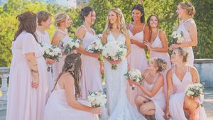A group wearing light lavender and white dresses gather together outdoors holding bouquets on a sunny day.