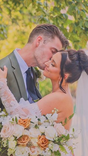 A romantic wedding couple embraces in a garden setting with peach and white roses in the foreground.