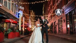 A couple in wedding attire shares a romantic moment on a city street illuminated by string lights at night.