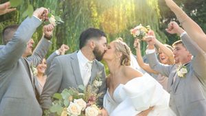 Joyful wedding party celebrating outdoors with raised bouquets against golden sunlight and willow trees.