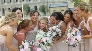 Bridesmaids in blush pink dresses share a joyful moment while holding pink and purple floral bouquets.