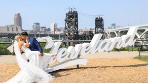 A couple poses for wedding photos in front of the Cleveland sign with the city skyline and bridges in the background.