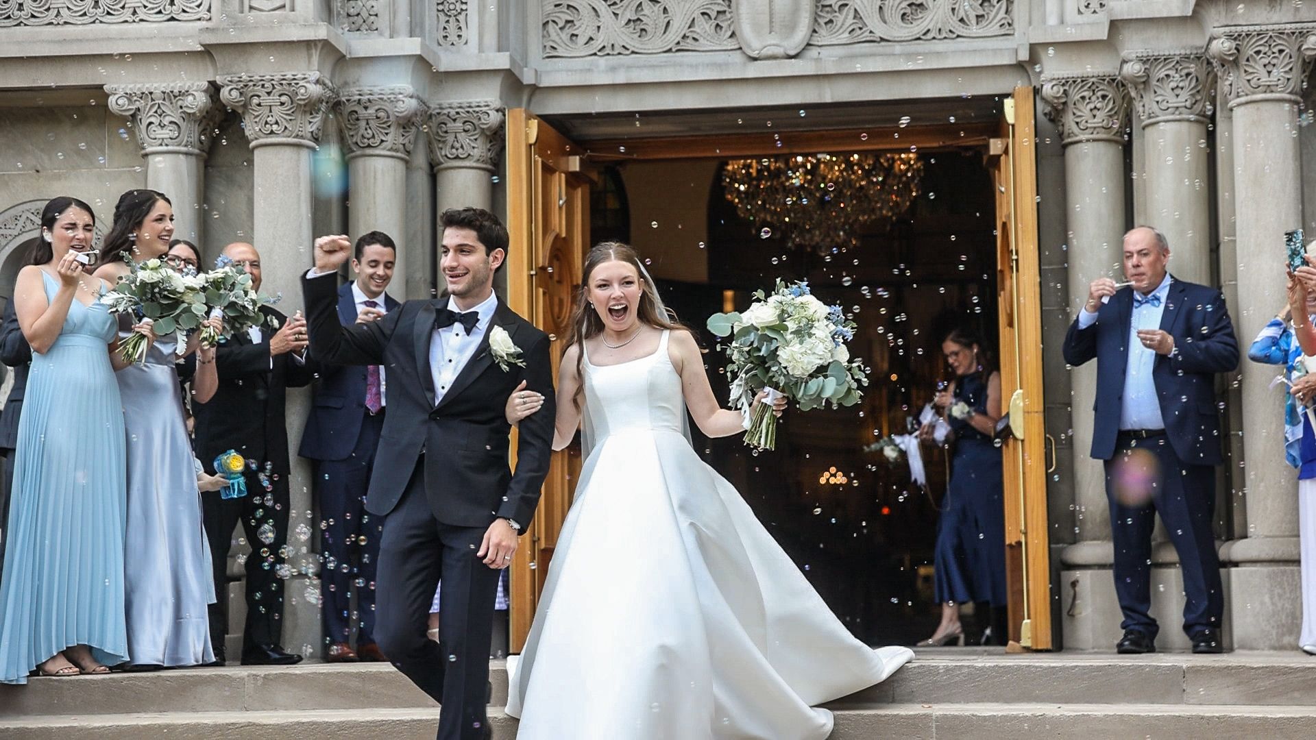 Newlyweds exiting ornate church doors while wedding guests celebrate with sparklers and flowers.