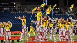 A marching band performs in striped uniforms with yellow flags at a nighttime stadium event.