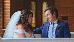 A couple shares a tender moment on their wedding day on a brick patio with arched windows in the background.