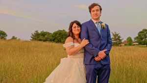 A couple in wedding attire stands together in a golden field at sunset with pine trees in the background.