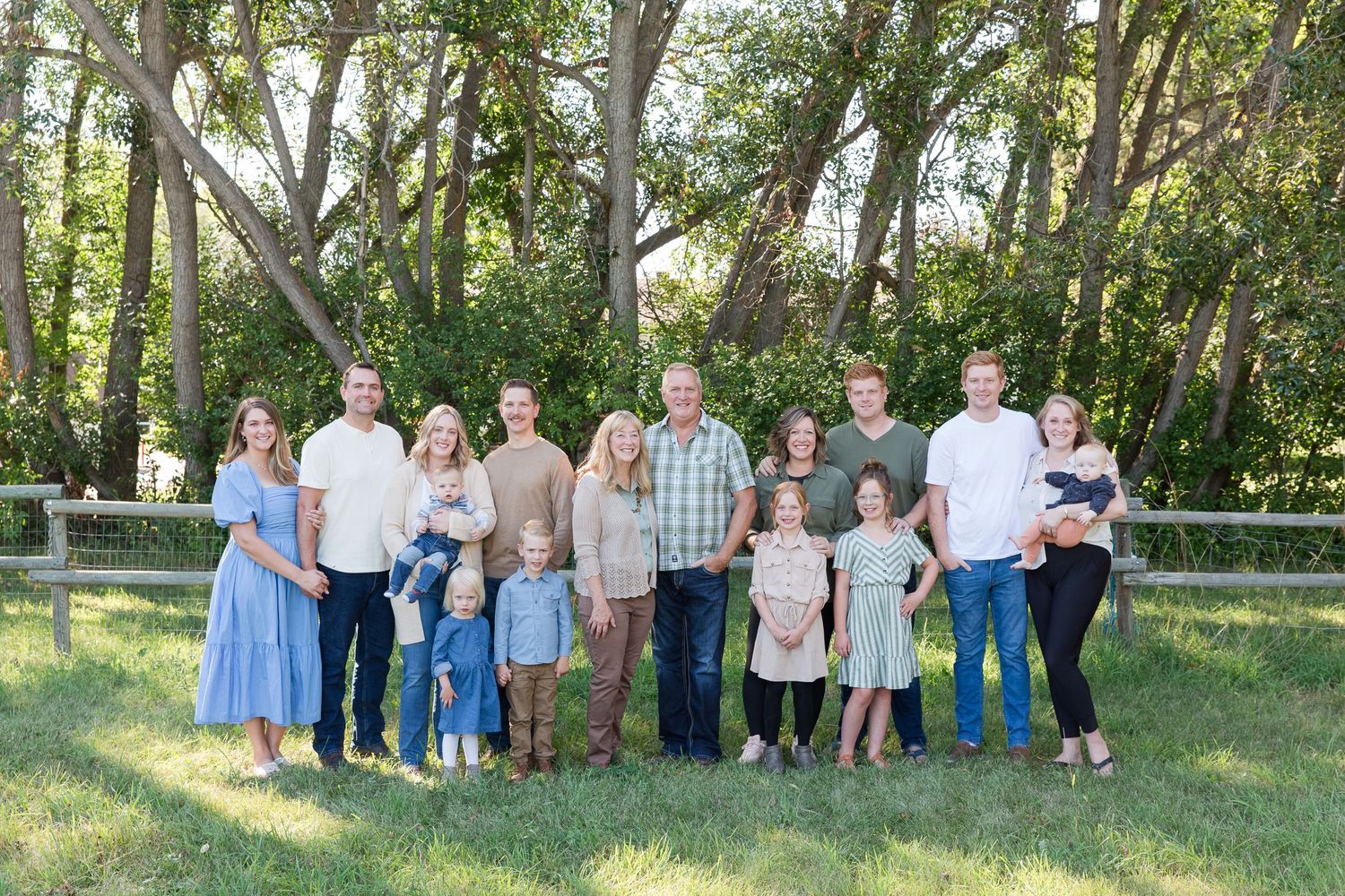 Extended family group portrait in casual attire gathered outdoors among green trees.