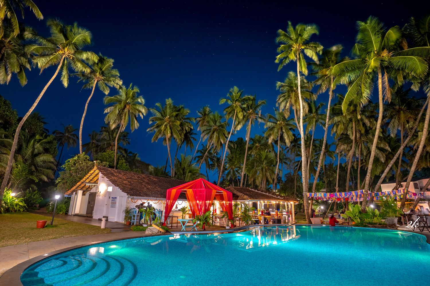 Tropical resort pool illuminated at night with palm trees and red tent structure.