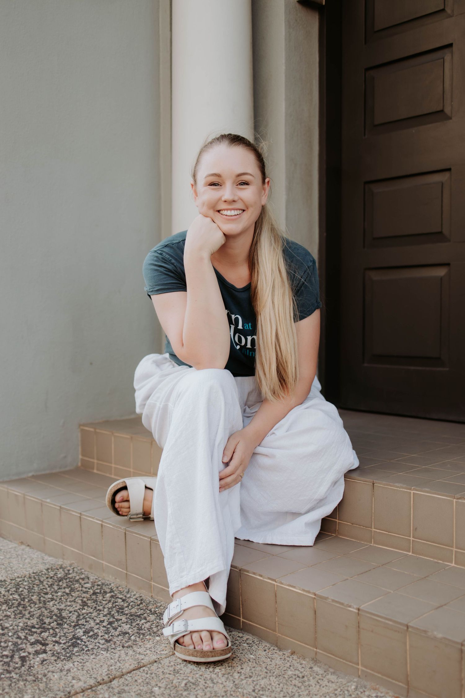 Sarah Del Borrello (Mother Mother Doula) in casual attire with white pants and sandals sits on concrete steps outside a building entrance.