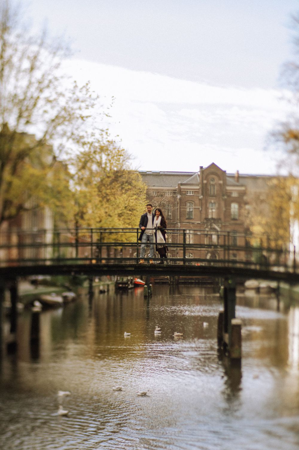 Panoramic view of a canal bridge in Amsterdam with autumn trees and historic buildings reflecting in the water.