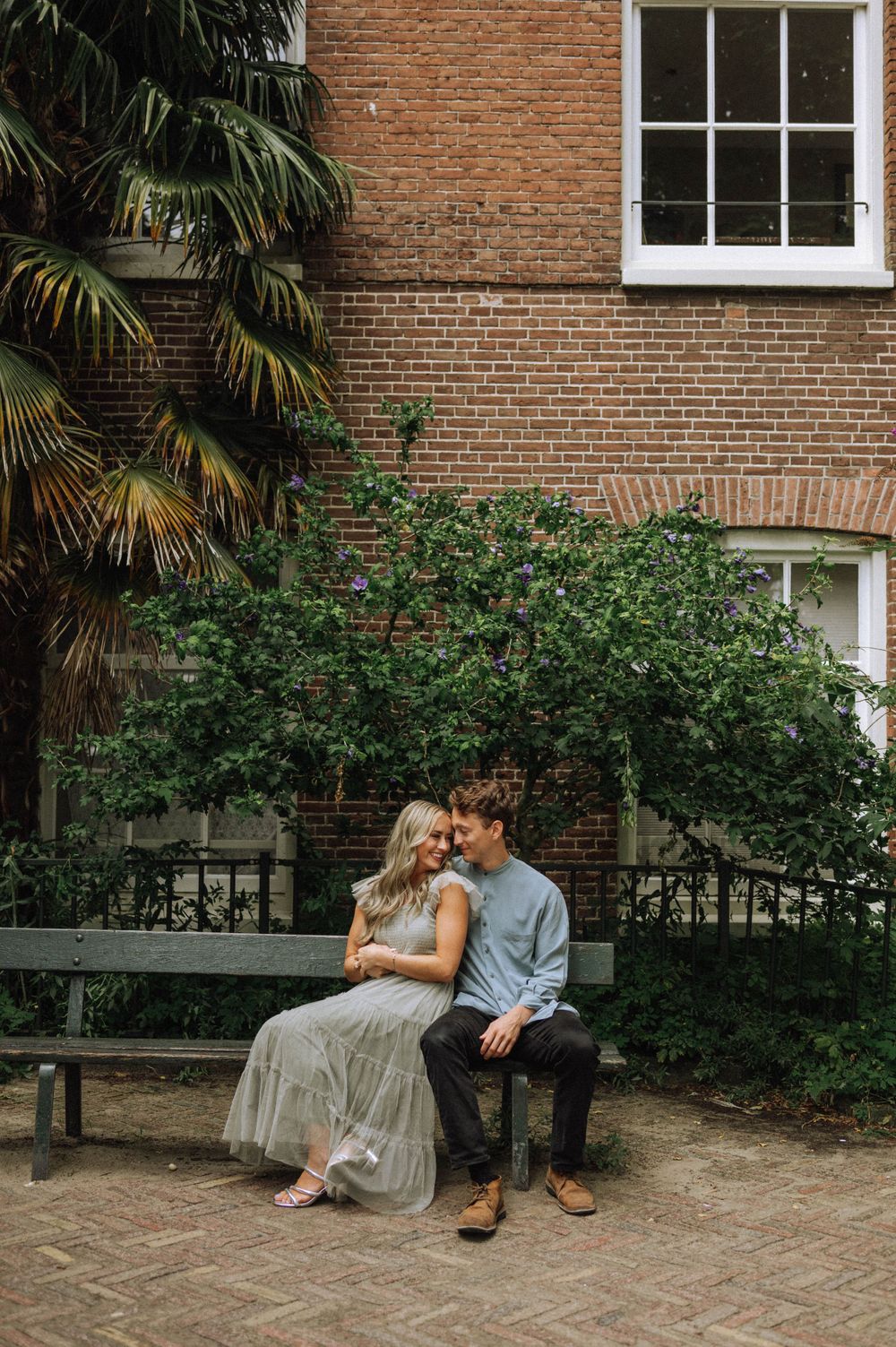 Couple sitting on a bench against brick wall with lush greenery and palm trees in an intimate moment.