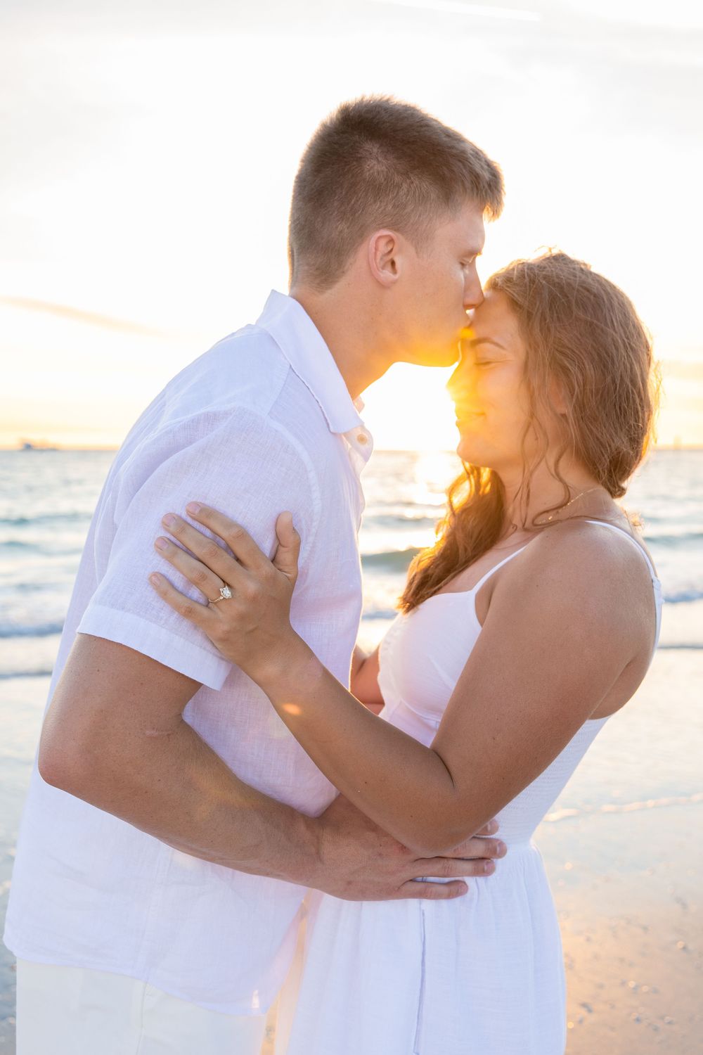 Romantic couple kissing on beach with golden sunset glowing behind them.
