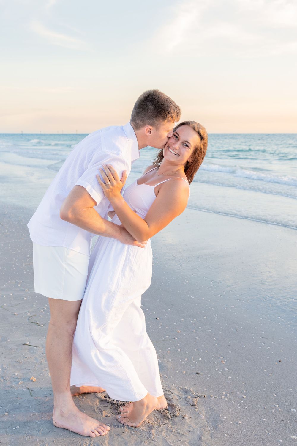 Couple embracing on beach at sunset, man kissing woman in white dress.