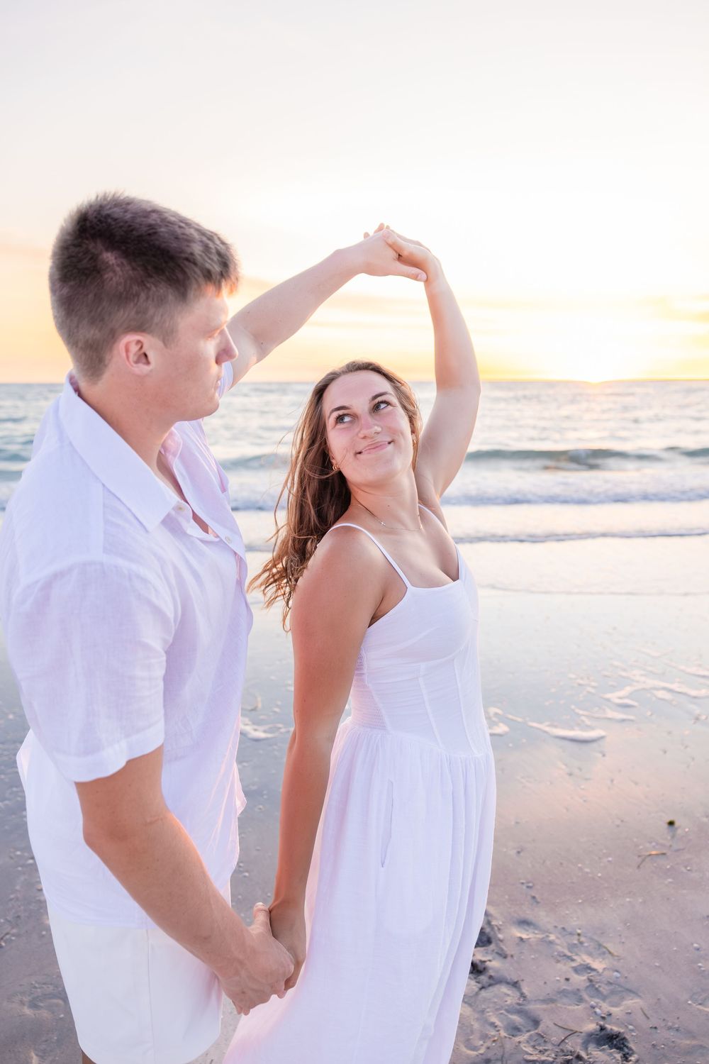 Young couple dancing on beach at sunset, woman in white dress spinning.