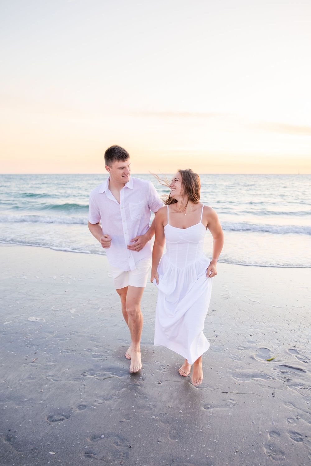 Happy couple in white outfits walking and laughing on beach at sunset.