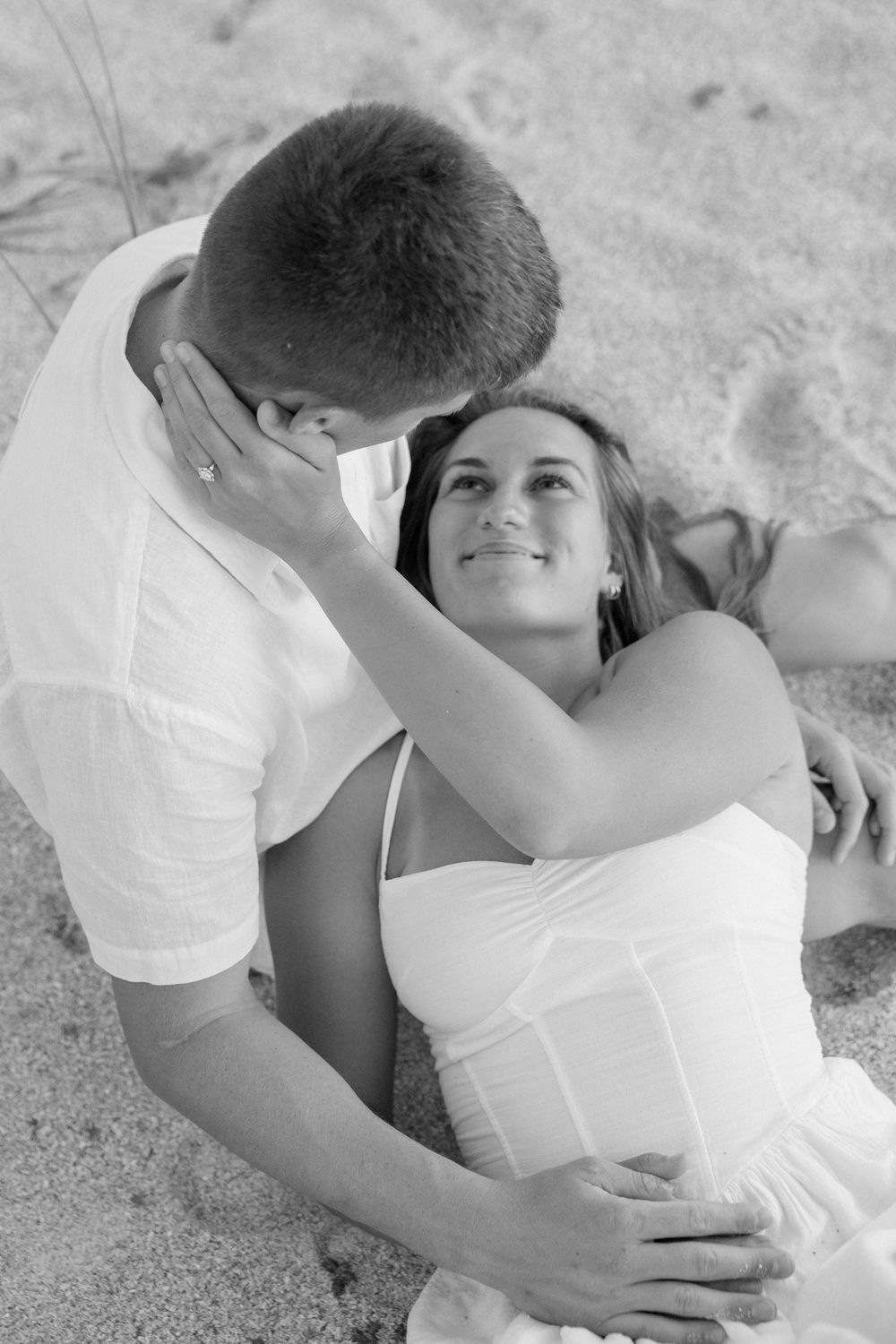 Black and white romantic beach photo of couple, woman in white dress smiling up at man while lying in sand.