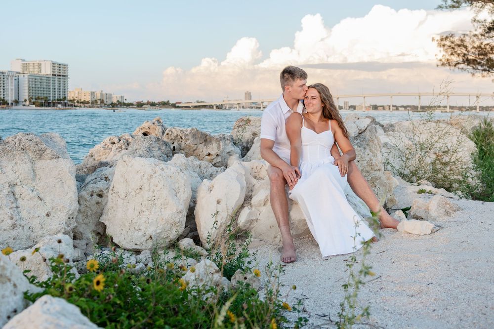 Couple sitting on rocky shoreline at sunset, woman in white dress, man in white shirt, with ocean and city skyline behind them.