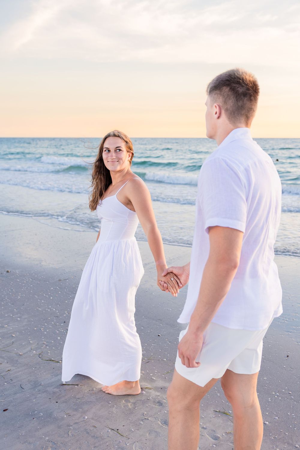 Young woman in white dress holding hands with man while walking on beach.
