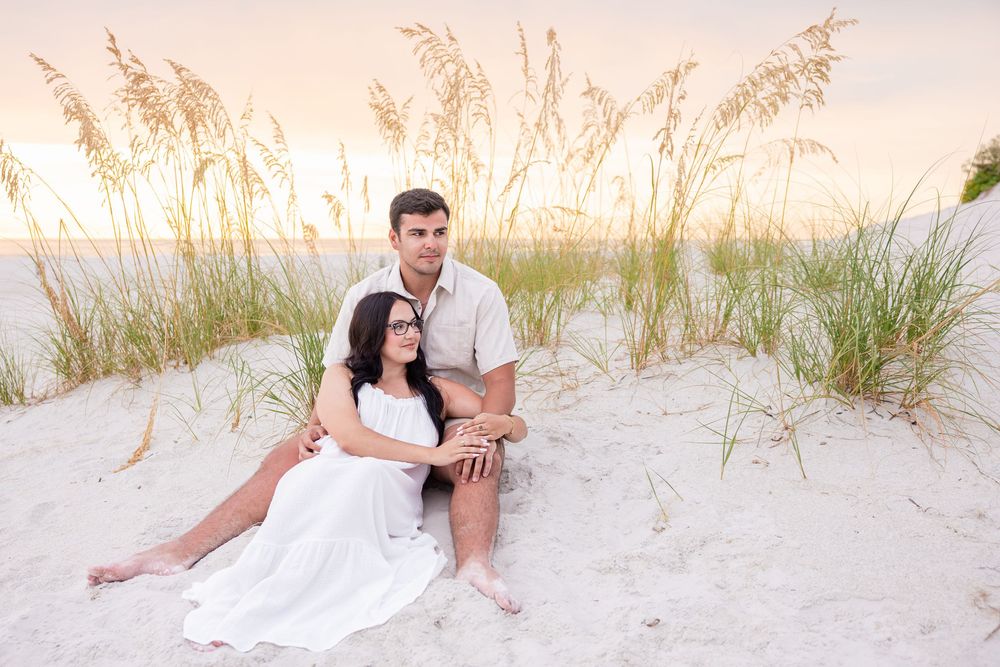 Couple sitting together on white sand dunes at sunset with sea oats, woman in white dress, man in casual attire.