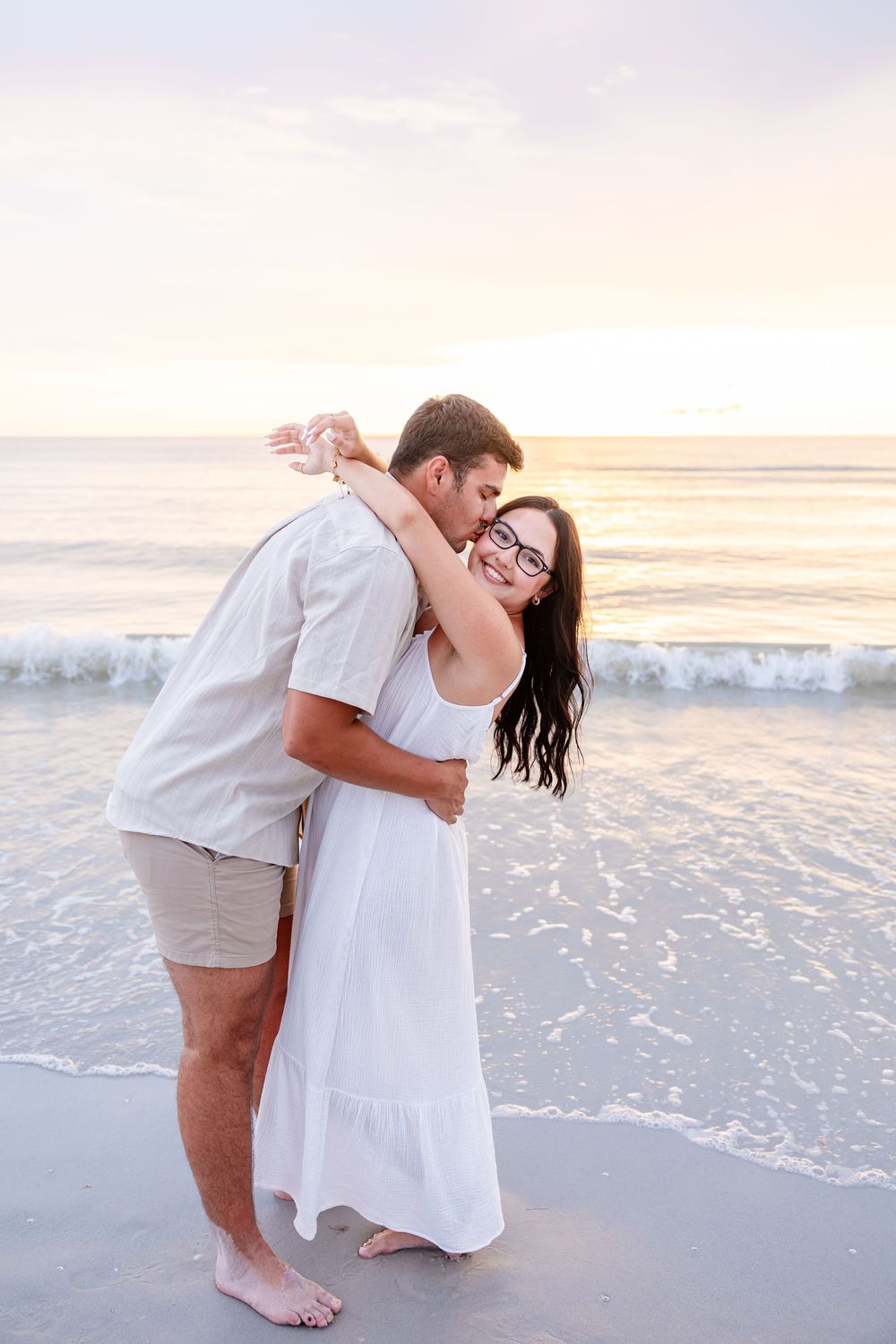 Couple embracing on a beach at sunset, woman in white dress smiling, man kissing her cheek, ocean waves behind them.