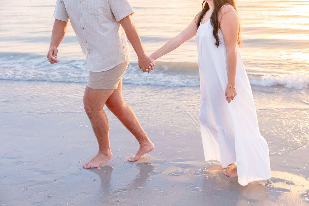 Couple holding hands walking barefoot on a sandy beach at sunset, woman in white dress, waves in background.