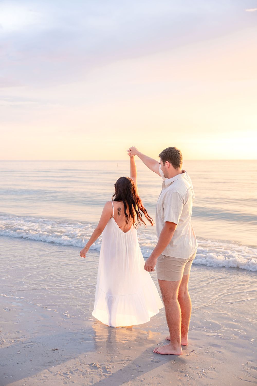 Couple dancing on beach at sunset, woman in white dress, man in khaki shorts, waves in background.