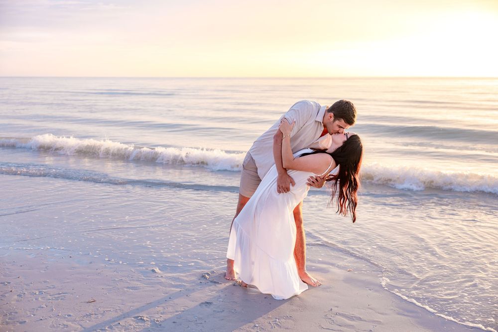 Romantic couple sharing a passionate kiss on a sandy beach at sunset, woman in white dress.