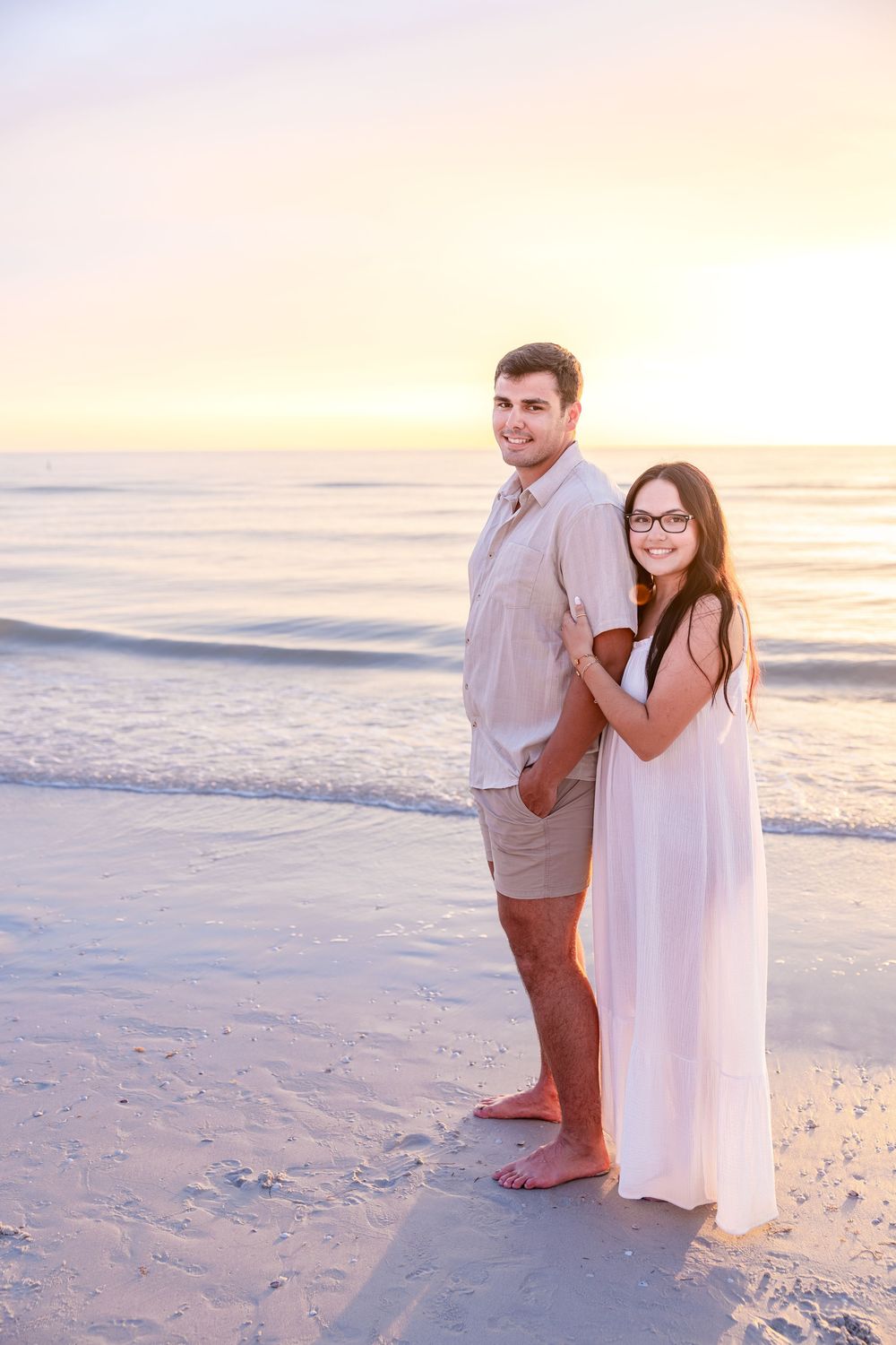 Couple embracing on a sandy beach at sunset, woman in white dress and man in khaki shorts smiling by the ocean.