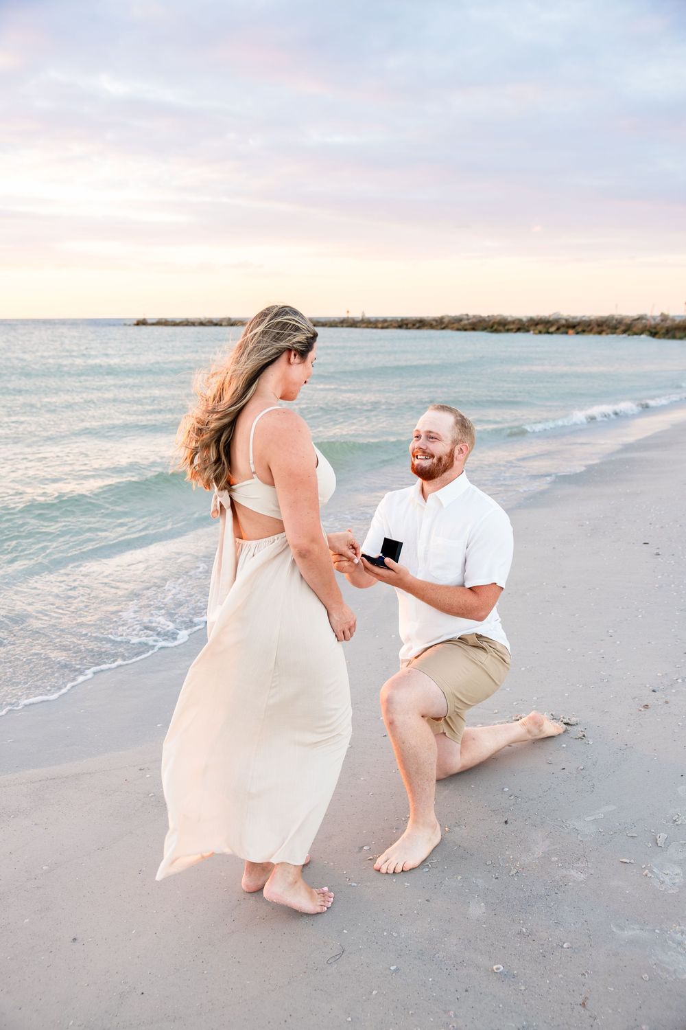 Man proposes on beach at sunset, kneeling with ring box while woman in white dress stands before him.