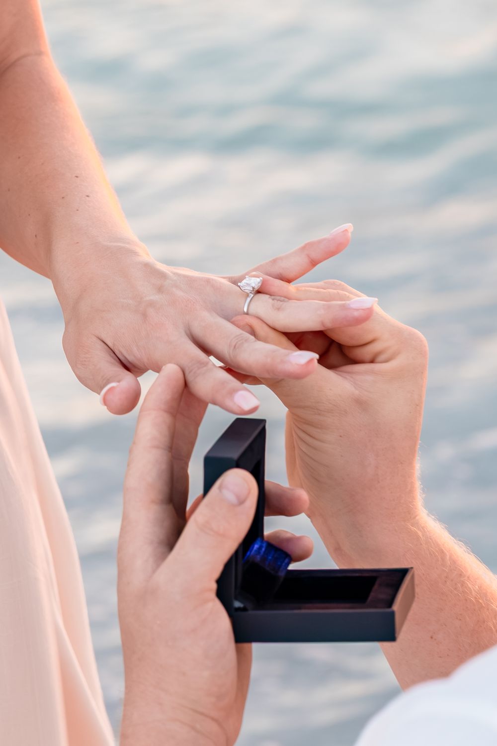 A man places a diamond engagement ring on a woman's finger during a romantic waterfront proposal.