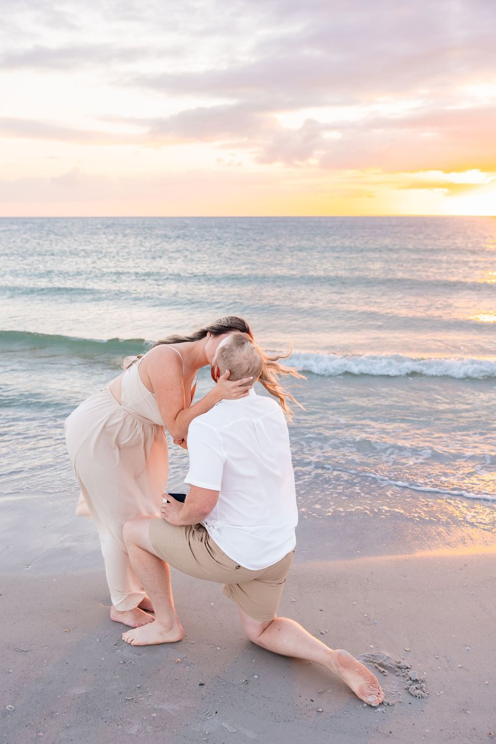 Man kneeling on beach proposing to woman at sunset with ocean waves in background.