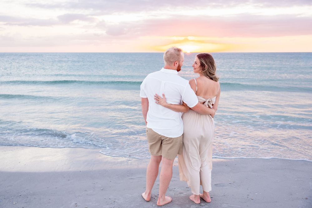Couple embracing on a sandy beach at sunset, gazing at each other with colorful sky and ocean waves behind them.