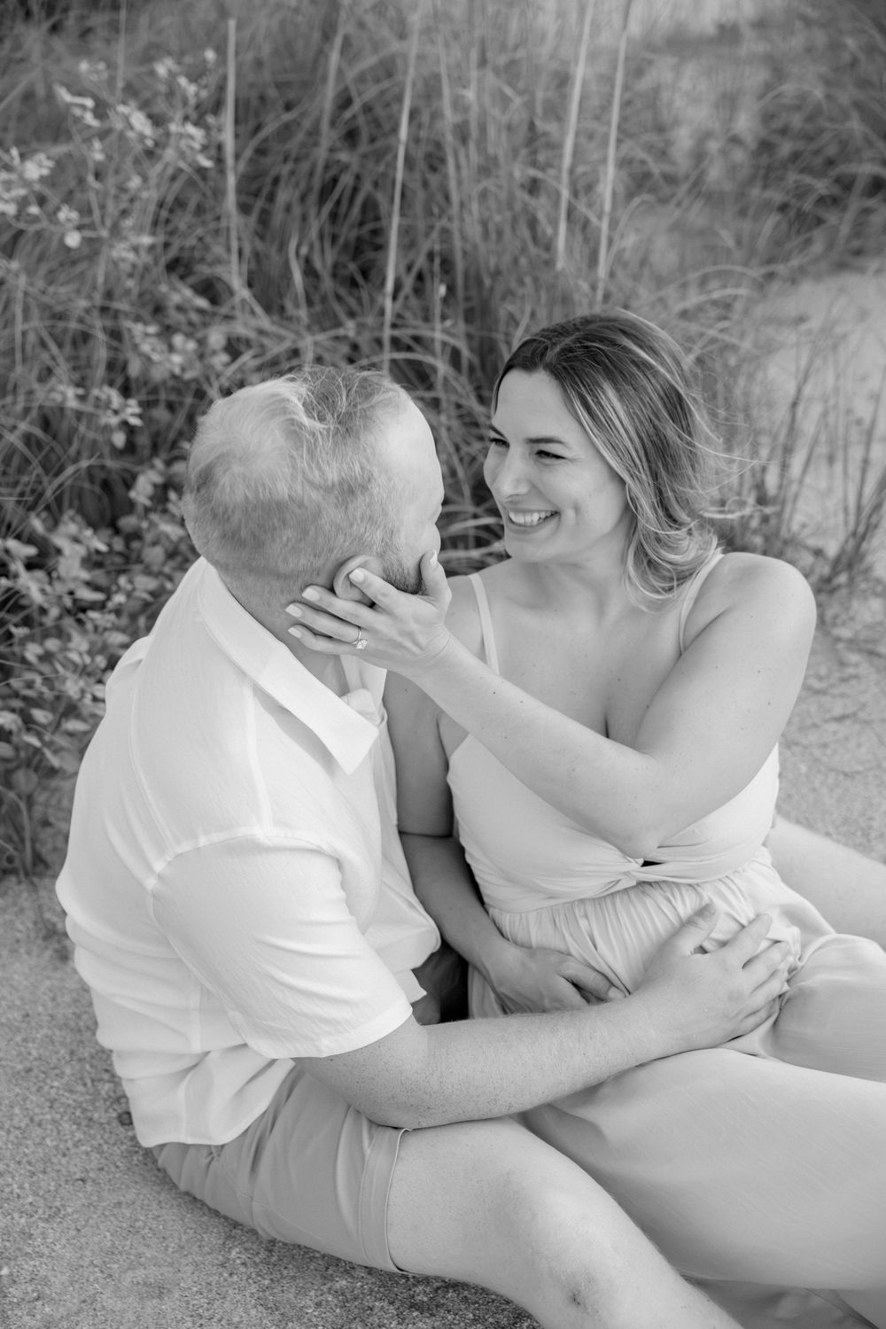 Black and white photo of a smiling couple sitting on sand, woman touching mans face, tall grass in background.