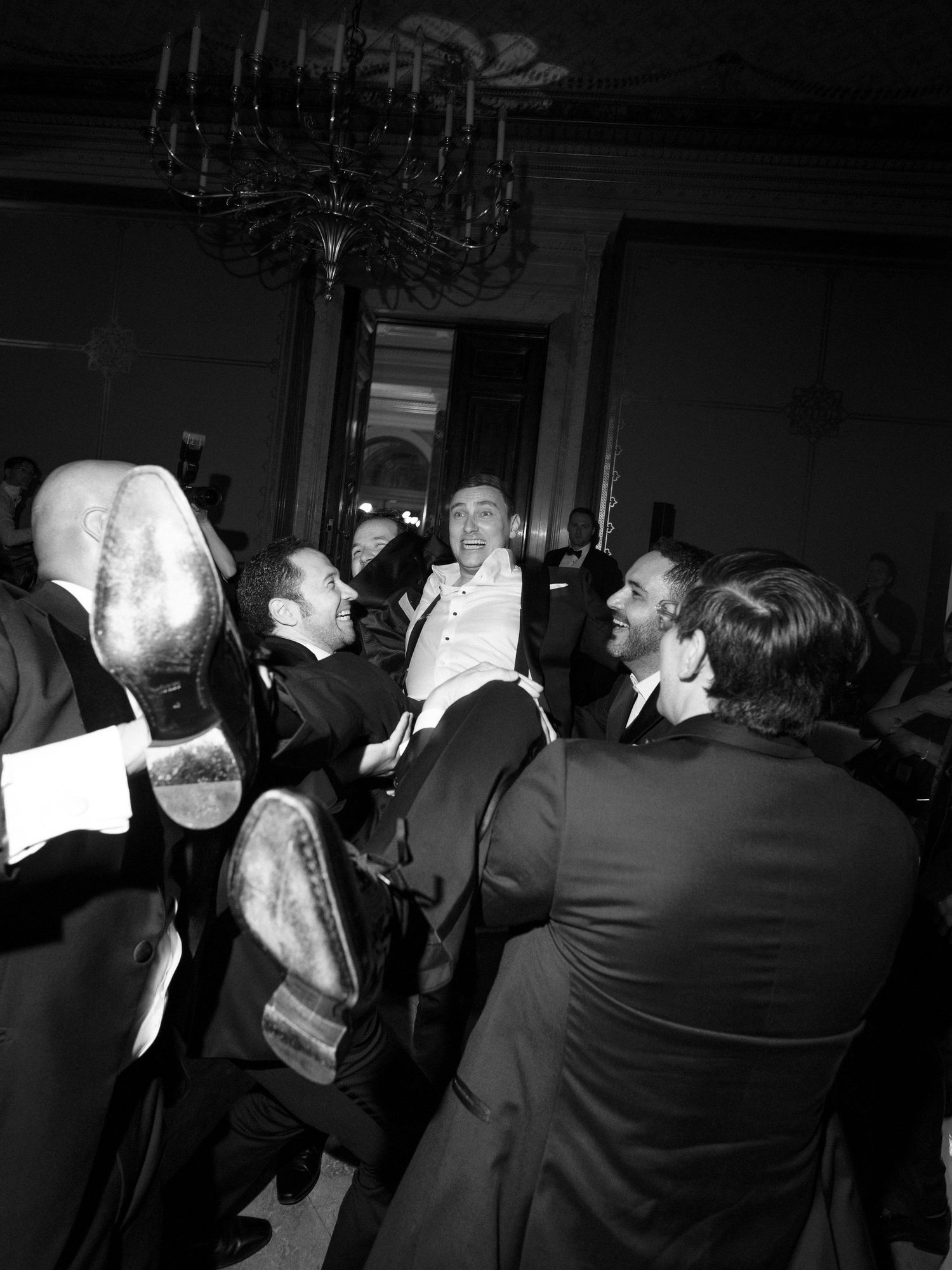 A black and white candid photo shows a lively group of men in formal attire laughing together under a chandelier.