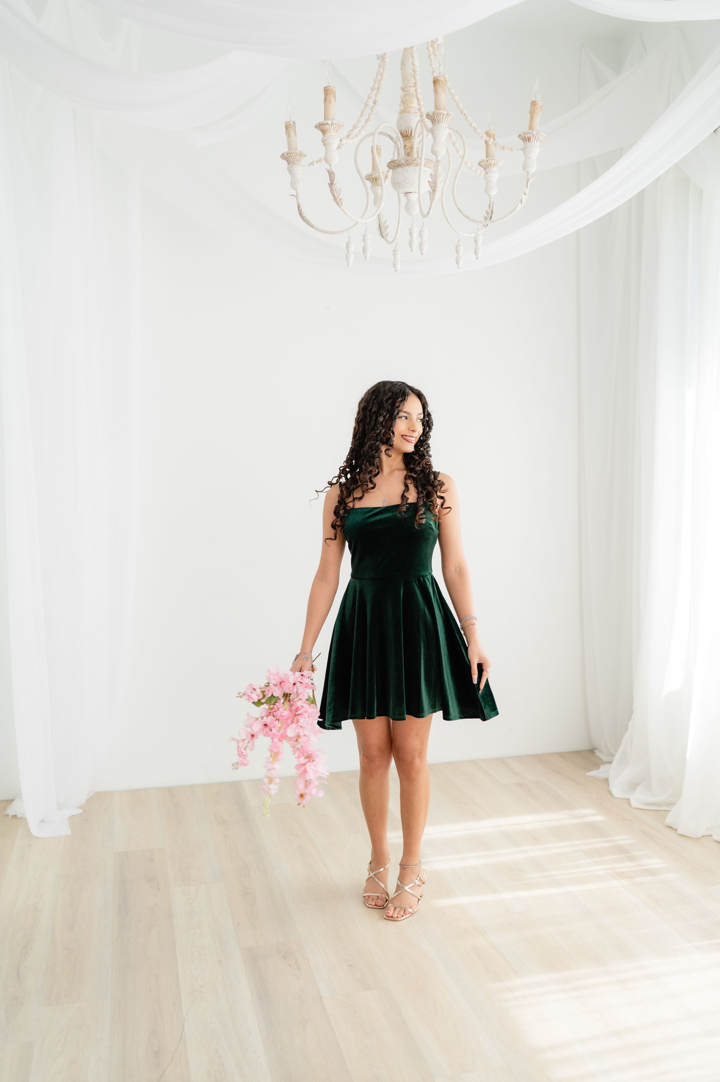 A woman in an elegant emerald green velvet dress poses in a bright white room with a chandelier and light hardwood floors.