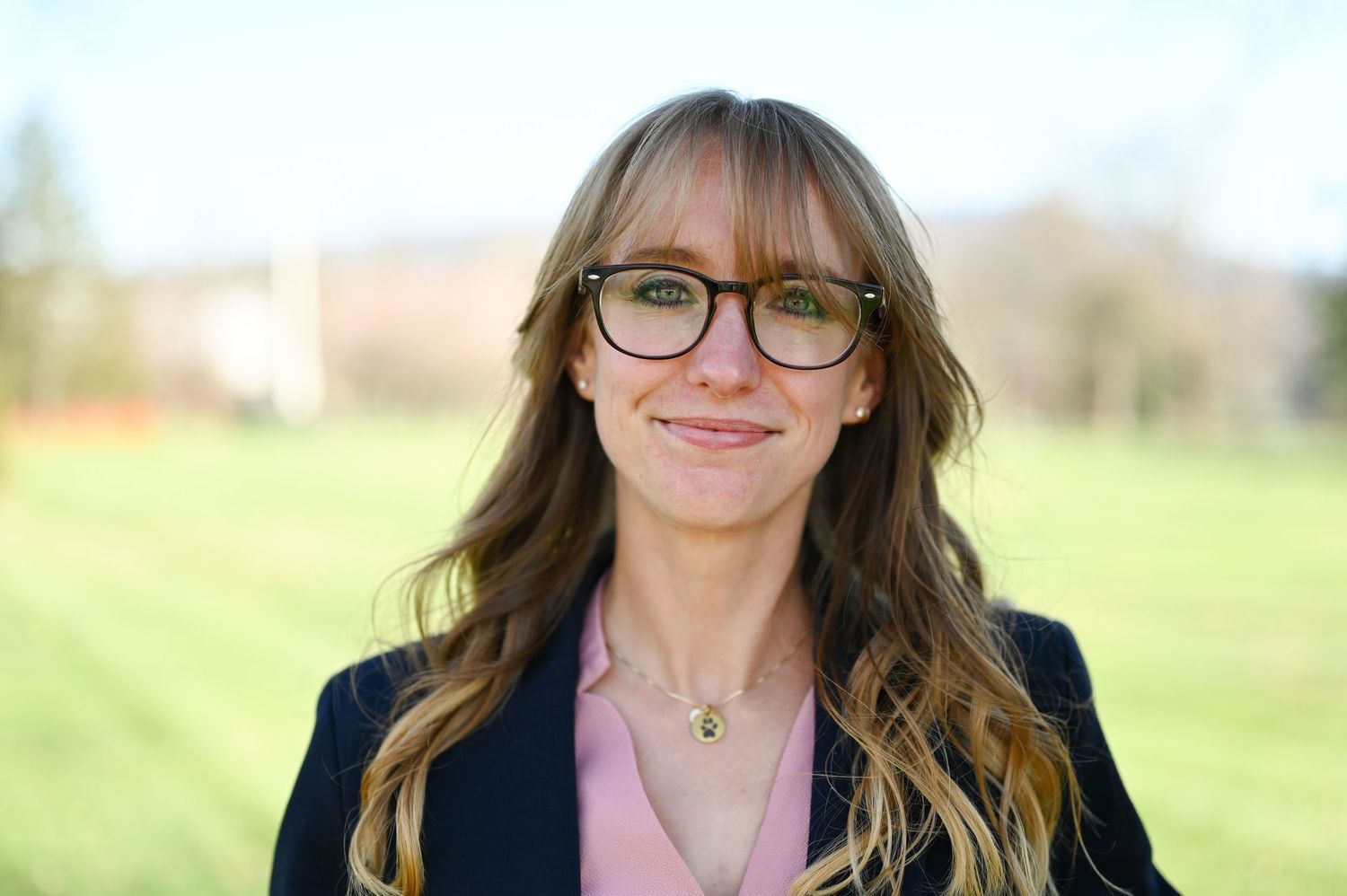Professional woman in glasses and blazer smiling outdoors on sunny day.