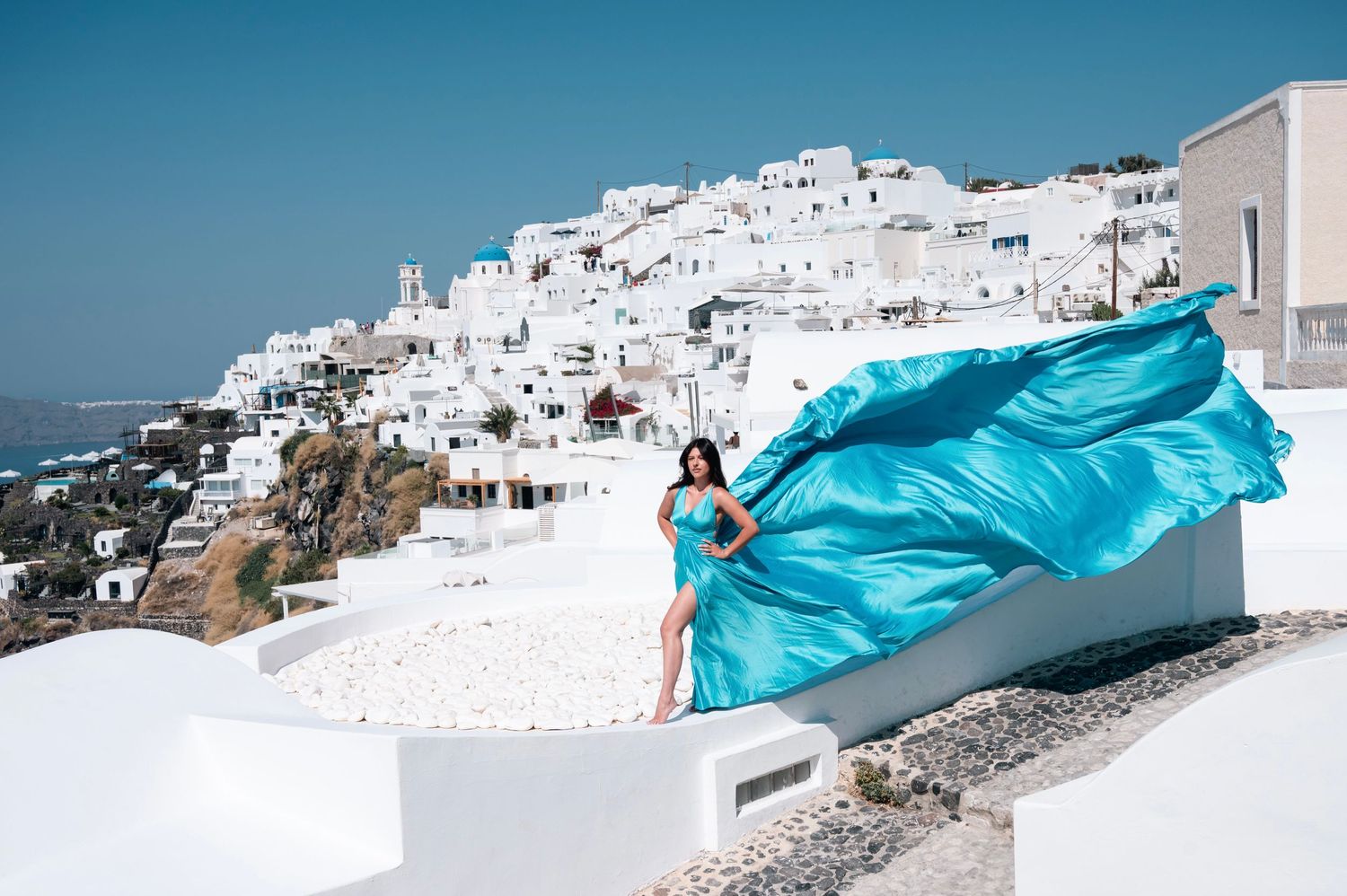 Woman in flowing turquoise dress poses against white buildings of Santorini, Greece.