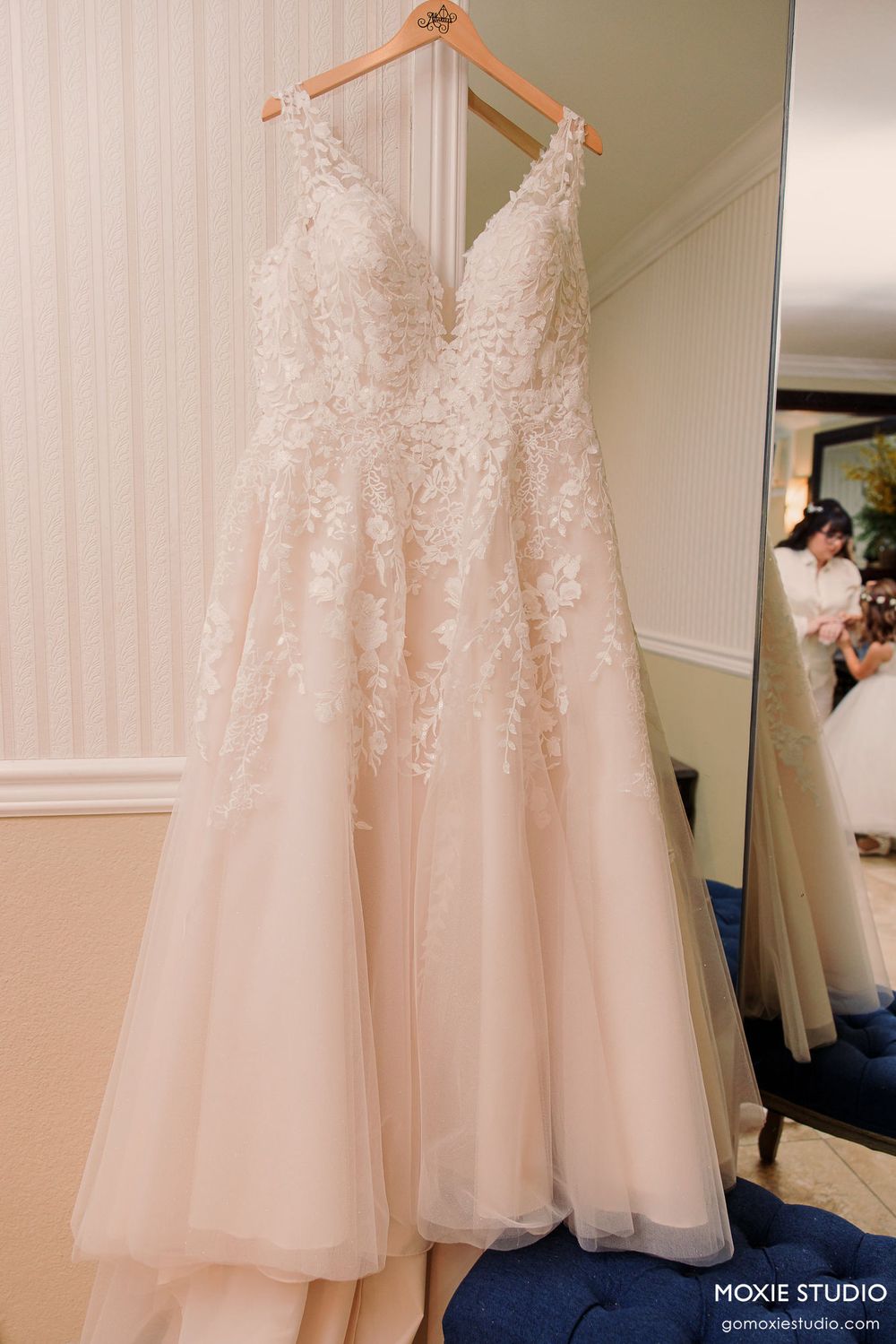 A delicate blush wedding gown with lace appliques and tulle skirt hangs on a wooden hanger against a cream wall.